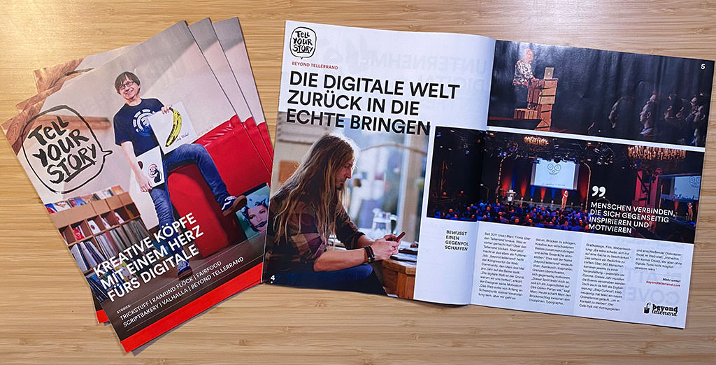 A photo showing a small pile of magazines on the left with its cover page and the title “Tell Your Story”. Next to it lies an open version on the right side with an interview of Marc Thiele about beyond tellerrand.