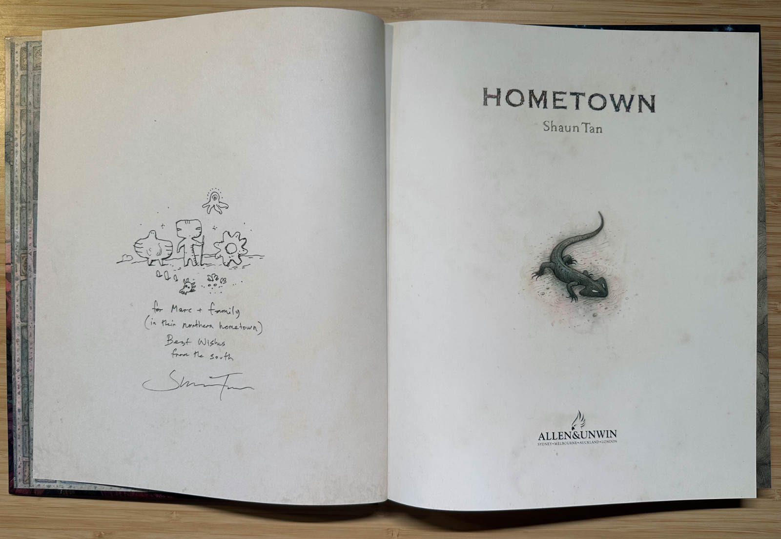 An open copy of Shaun Tan&rsquo;s &ldquo;Hometown&rdquo;. The left page has a small ink drawing of odd little creatures standing together, a handwritten dedication and Shaun&rsquo;s signature. The right page shows the title page with a small drawing of a lizard.