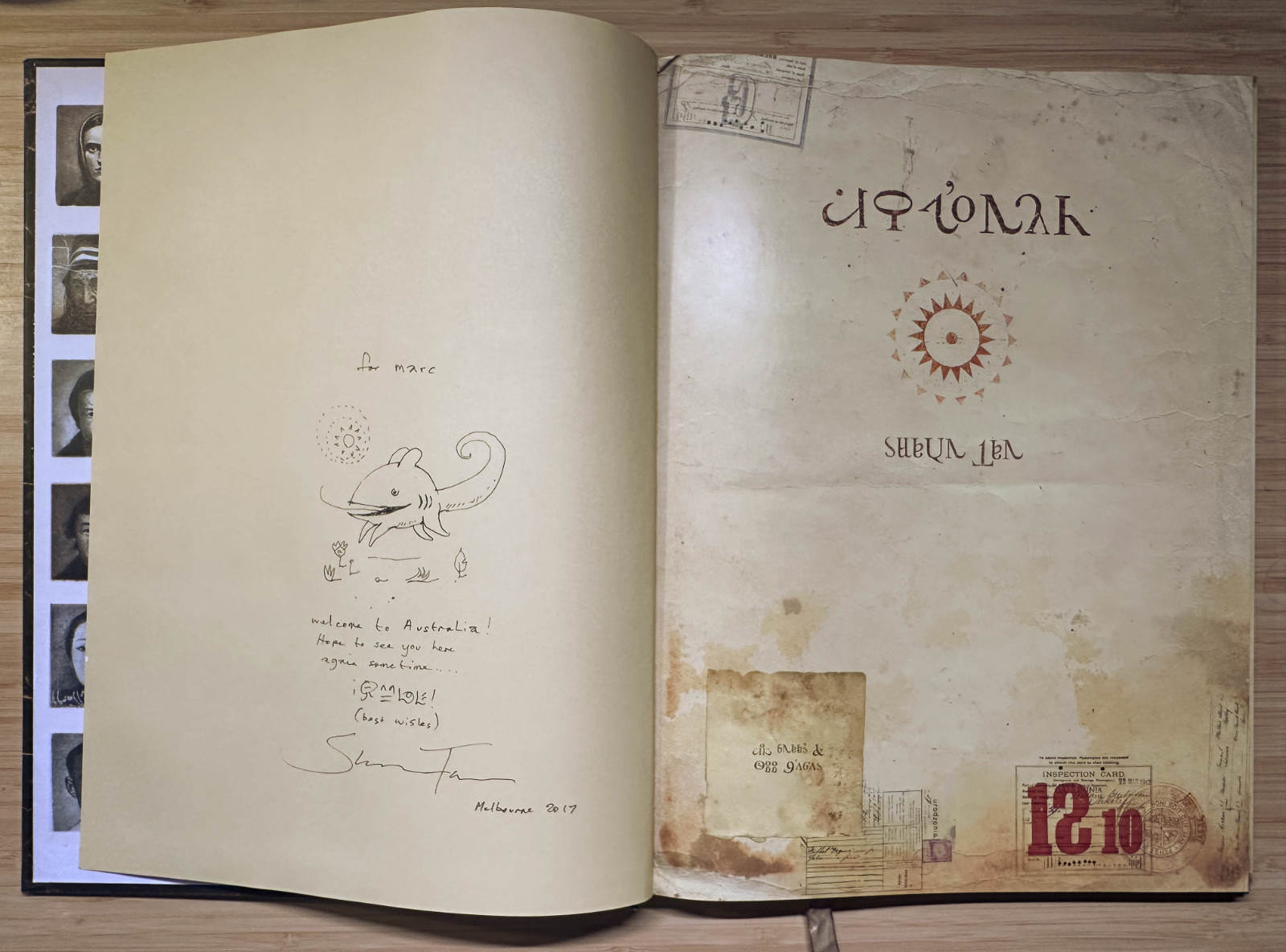 An open copy of Shaun Tan&rsquo;s &ldquo;The Arrival&rdquo;. The left page has an ink drawing of a small grinning creature with a curled tail under a dotted sun. Around it Shaun wrote &ldquo;for marc&rdquo;, &ldquo;Hope to see you here again sometime &hellip;&rdquo; and &ldquo;best wishes&rdquo; in the book&rsquo;s invented alphabet, signed and dated &ldquo;Melbourne 2017&rdquo;. The right page shows the aged title page.