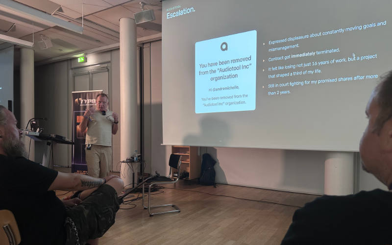 Andr&eacute; Michelle speaks and gestures on stage at TYPO3 Developer Days 2026. The projection screen behind him, titled &lsquo;Audiotool Escalation&rsquo; shows a GitHub notification reading &lsquo;You have been removed from the Audiotool Inc organization&rsquo; alongside bullet points describing his displeasure with mismanagement, the immediate termination of his contract, the emotional impact of losing 16 years of work and an ongoing court case over promised shares. Audience members are visible in the foreground.