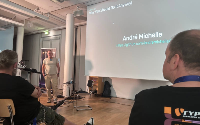 Andr&eacute; Michelle stands in front of a large projection screen at TYPO3 Developer Days 2026, presenting a title slide that reads &lsquo;Why You Should Do It Anyway!&rsquo; along with his name and GitHub URL. Two audience members are visible in the foreground, one wearing a TYPO3 t-shirt.