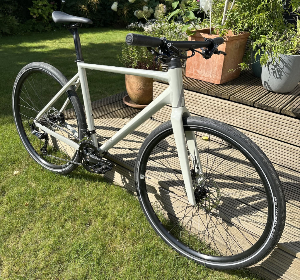 A photo of my new bike, shot in my garden. You see a light grey urban bike with black tires leaning against a wooden terrace, standing on green lawn.