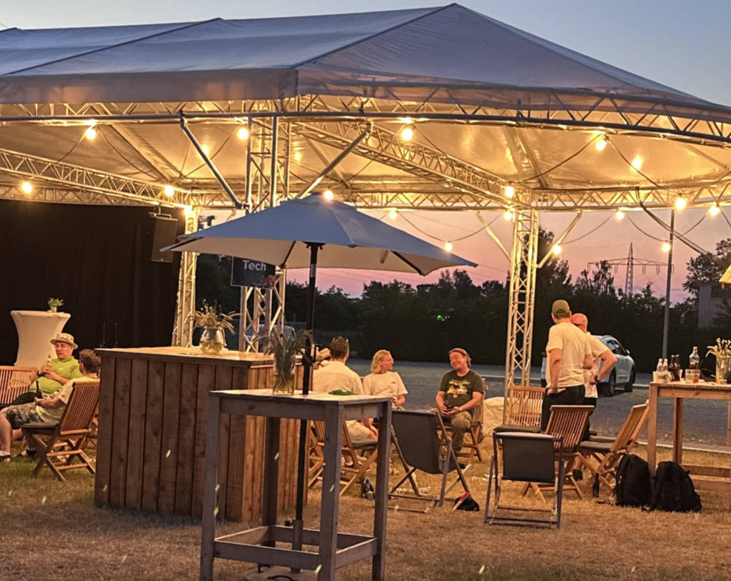 Outdoor evening scene at the Head in the Cloud event in Espelkamp. A large open-sided tent strung with warm fairy lights shelters a casual chill-out area with wooden bar furniture, folding chairs and a parasol. Small groups of attendees sit and stand around, chatting and having drinks, with a dusk sky and trees in the background.