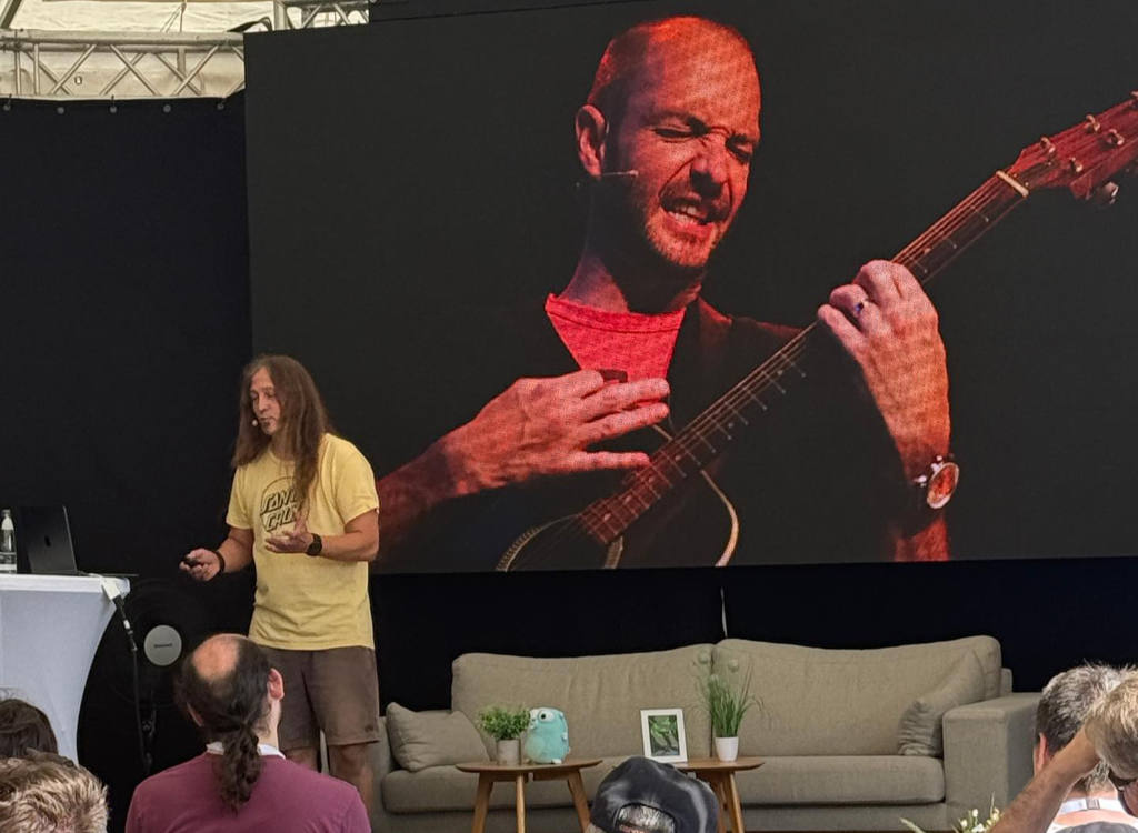 Marc Thiele stands on stage at Head in the Cloud in Espelkamp, wearing a yellow t-shirt and headset microphone, gesturing as he speaks. Behind him, a large screen shows a close-up of Josh Brewer passionately playing a quitar. A beige sofa with small plants and picture frames sits at the front of the stage, with audience members visible in the foreground.