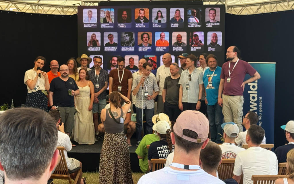 Group photo on stage at Head in the Cloud in Espelkamp. A large group of speakers and organisers stand together, smiling and chatting, in front of a backdrop showing portraits and names of the event&rsquo;s speakers. The audience is visible in the foreground, watching from their seats. A 'mittwald' banner stands to the right of the stage.