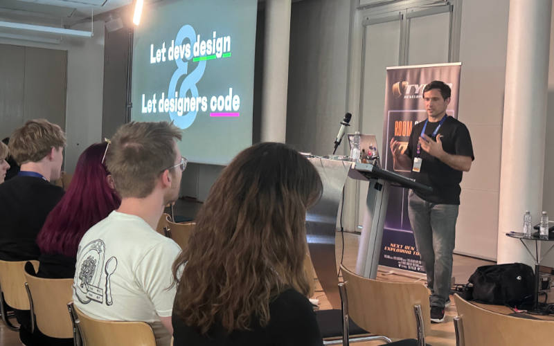 Matthias Ott stands at a lectern speaking to an audience at TYPO3 Developer Days Karlsruhe 2026. A projected slide on the left reads &lsquo;Let devs design & Let designers code&rsquo; with &lsquo;design&rsquo; underlined in green and &lsquo;code&rsquo; underlined in pink. A TYPO3 &lsquo;Room Sponsor&rsquo; banner stands next to the lectern, and attendees are seated on wooden chairs in the foreground.