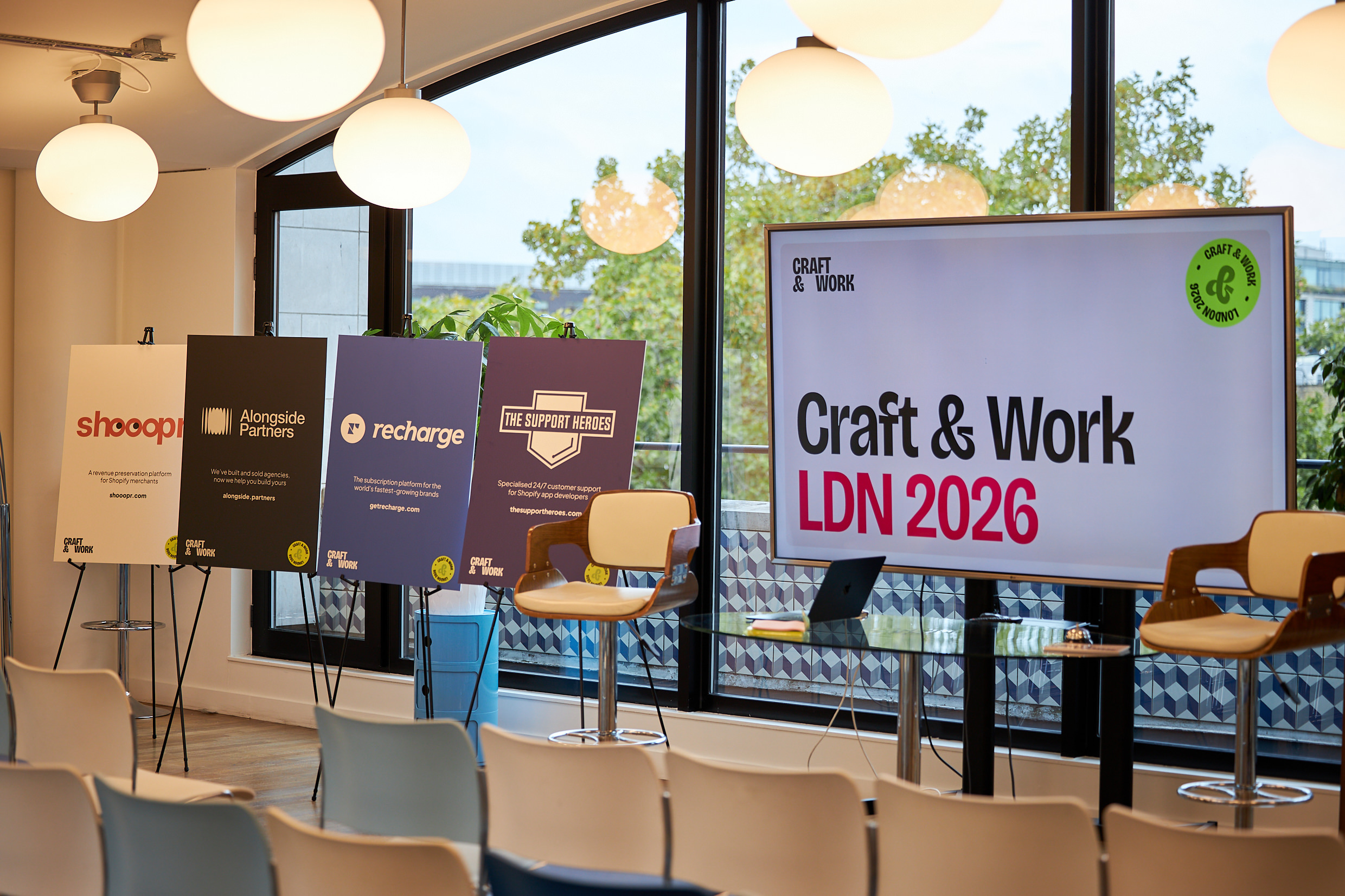 Interior view of the Craft & Work LDN 2026 event space in London. A large screen on the right displays the event logo &lsquo;Craft & Work LDN 2026&rsquo; in bold black and pink type. On the left, four sponsor posters on easels showcase Shooopr, Alongside Partners, Recharge, and The Support Heroes. Two bar stools and a glass table with a laptop sit on stage in front of a large window with trees visible outside, and rows of empty chairs face the stage. Spherical pendant lights hang from the ceiling.