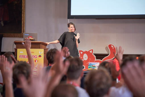 A speaker with long hair stands on stage at SmashingConf Freiburg 2026, smiling and waving as audience members raise their hands in the foreground. A red 'Freiburg' Smashing Cat mascot cutout stands behind them next to the lectern.
