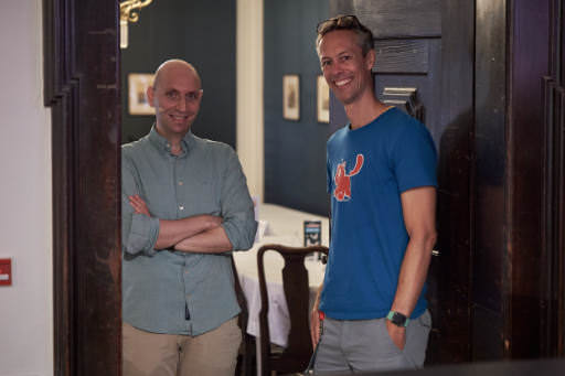 Two men stand smiling in a doorway at SmashingConf Freiburg 2026. On the left, a man with a shaved head wears a blue-grey shirt with arms crossed. On the right, a taller man wears a blue t-shirt with a red Smashing Cat logo.