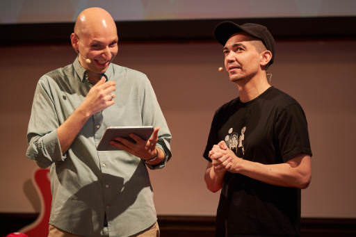 Two people stand on stage at SmashingConf Freiburg 2026, chatting and smiling. Left is Vitaly Friedman, right is Heydon Pickering. Vitaly wears a light blue shirt and holds a tablet, while Heydon wears a black cap and t-shirt. Both have headset microphones.