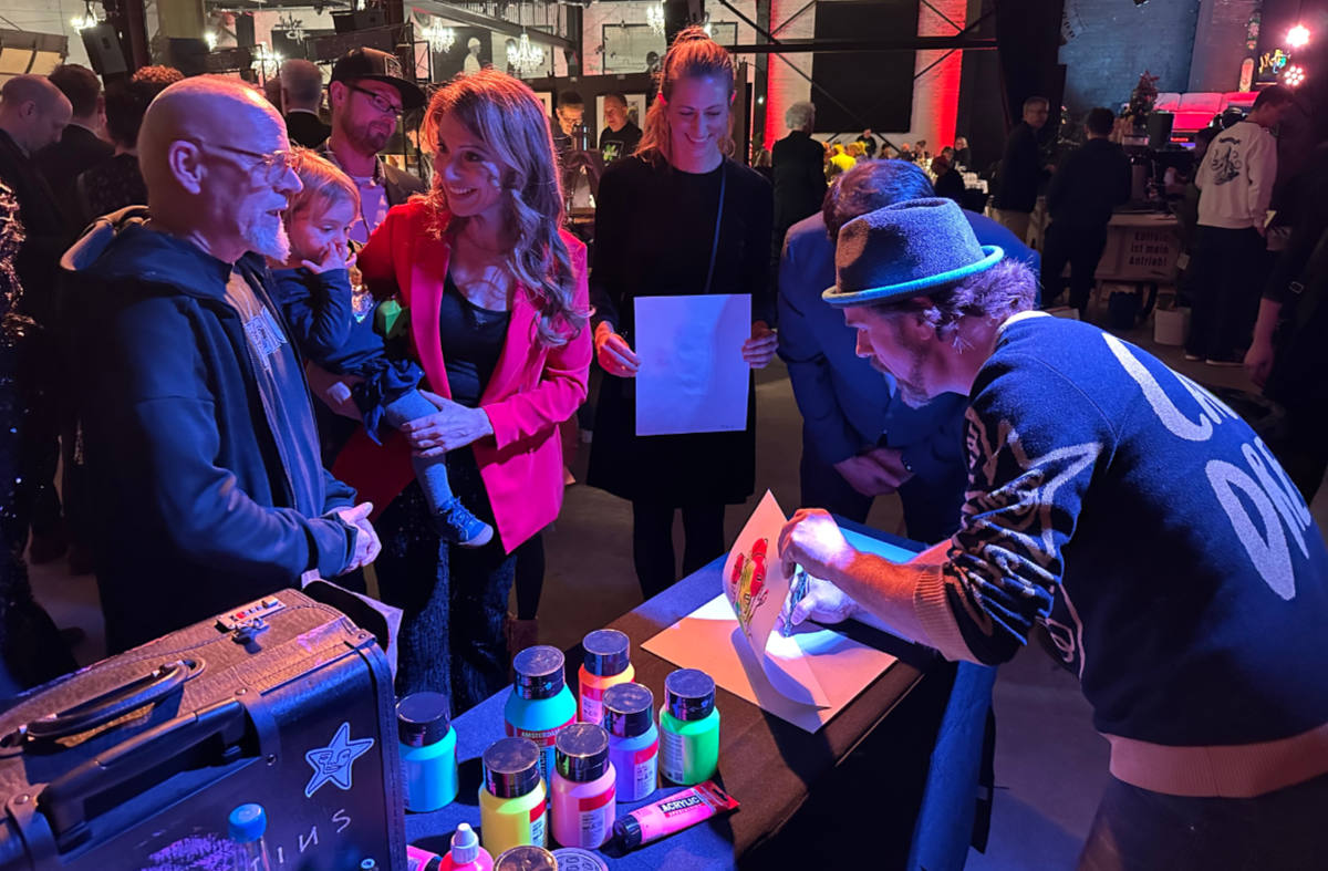 A table surrounded by people. You see Thomas D and Mara Bergmann amongst the people watching the artists Salventius drawing.