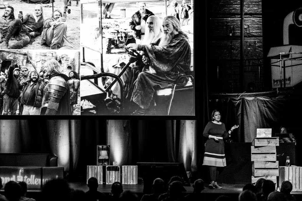 Black-and-white photo of Sacha Judd on stage at beyond tellerrand Düsseldorf 2022. She stands on the right in a dress, gesturing as she speaks to the audience. A large screen behind her displays behind-the-scenes photos from a Lord of the Rings film shoot, including Ian McKellen as Gandalf in costume driving a vehicle and cast members with director Peter Jackson on set. Stacked wooden crates and stage lamps frame the scene, with silhouettes of the audience visible in the foreground.